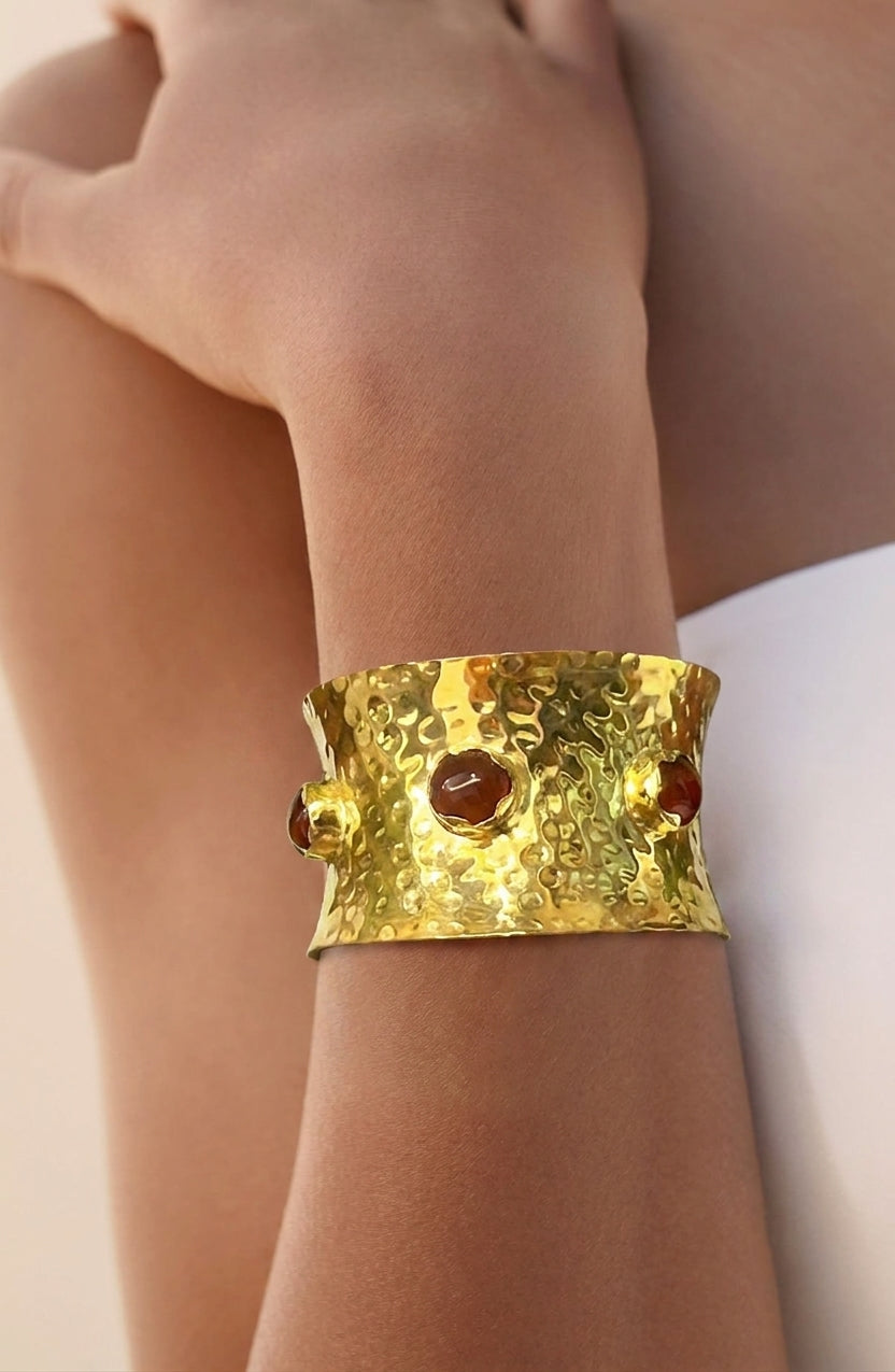 A close-up of a person's wrist wearing a wide, hammered gold cuff bracelet with three round amber-brown Carnelian gemstones.
