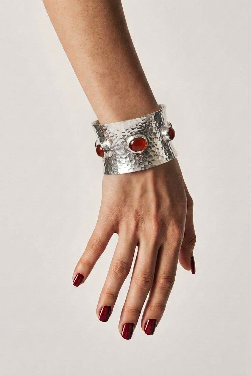 A silver hammered cuff bracelet with three red Carnelian gemstones, shown worn on a person's wrist with dark red nail polish against a light gray background.