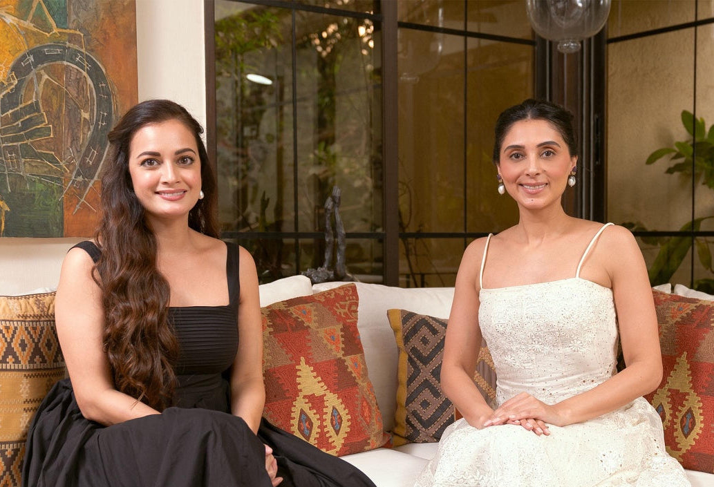 Two women seated  on a white sofa in a modern living room setting. The woman on the left wears a long black sleeveless gown, and the woman on the right wears a white sleeveless lace dress.