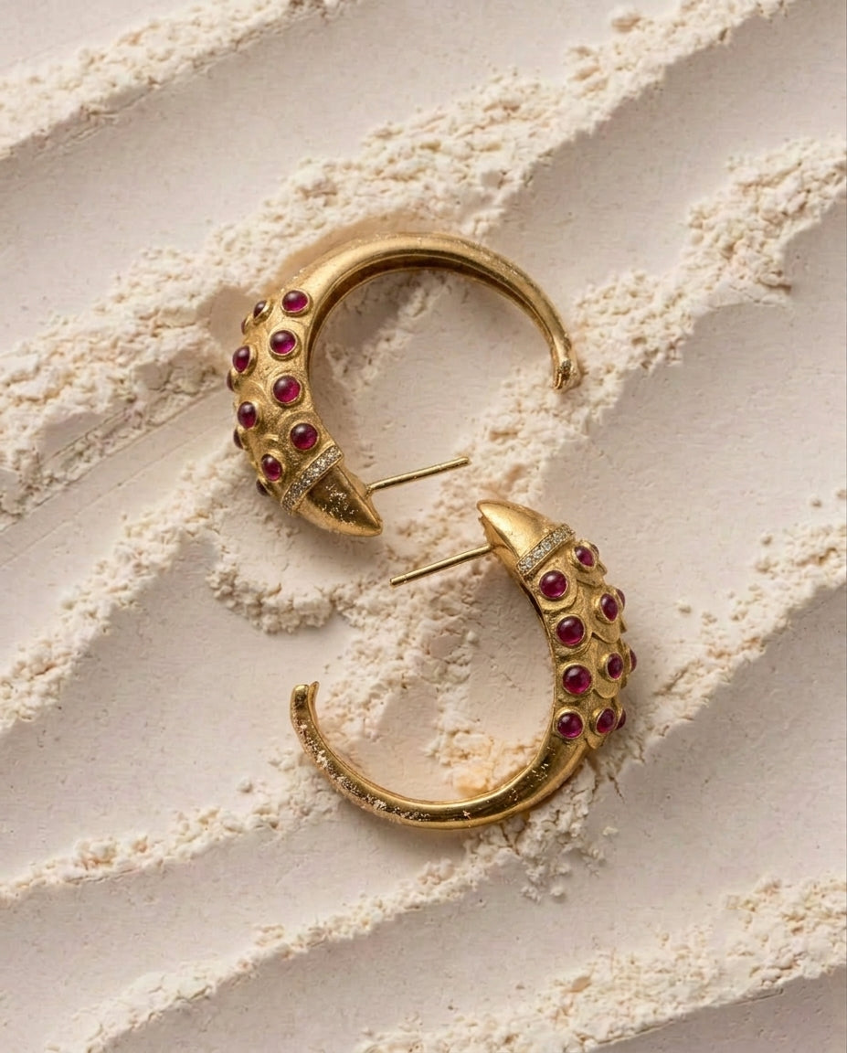 A pair of gold crescent-shaped hoop earrings embellished with small Ruby Red Cabochons gemstones, arranged on a textured beige surface.