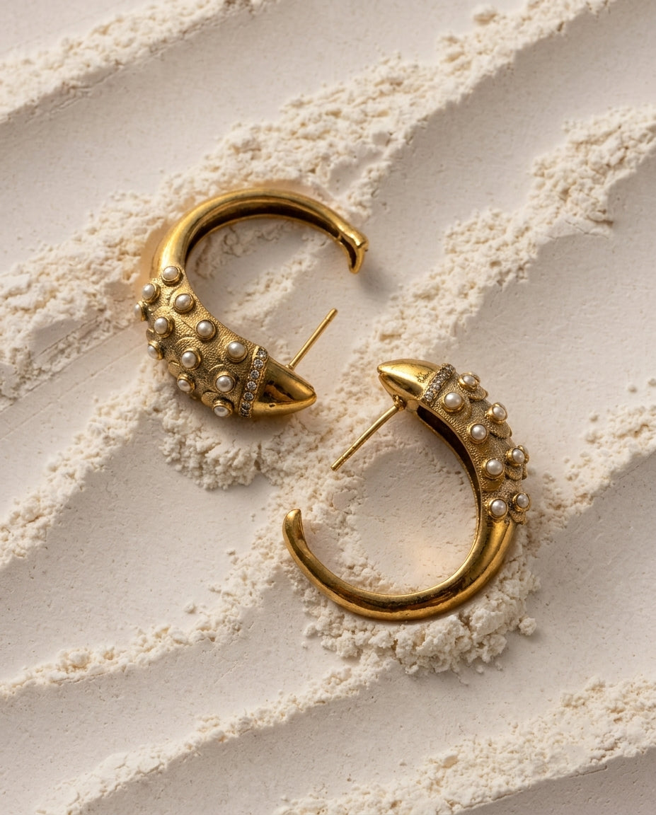 A pair of gold crescent-shaped hoop earrings embellished with small Freshwater pearl-like beads and clear rhinestones, arranged on a textured beige surface with fine powder.