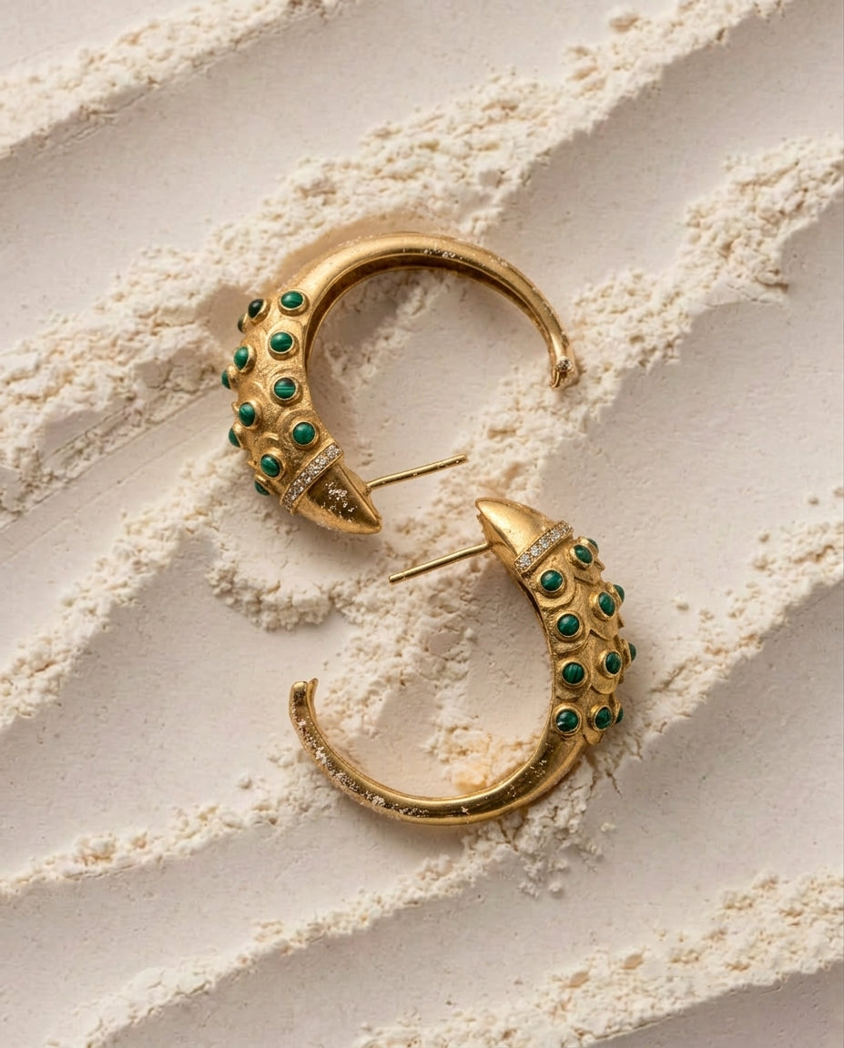 A pair of gold crescent-shaped hoop earrings embellished with small green Malachite Cabochons stones, arranged on a textured beige surface.