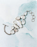 A silver-toned statement bracelet featuring irregular, organic-shaped white mother-of-pearl stones, displayed on a textured light blue and white background.