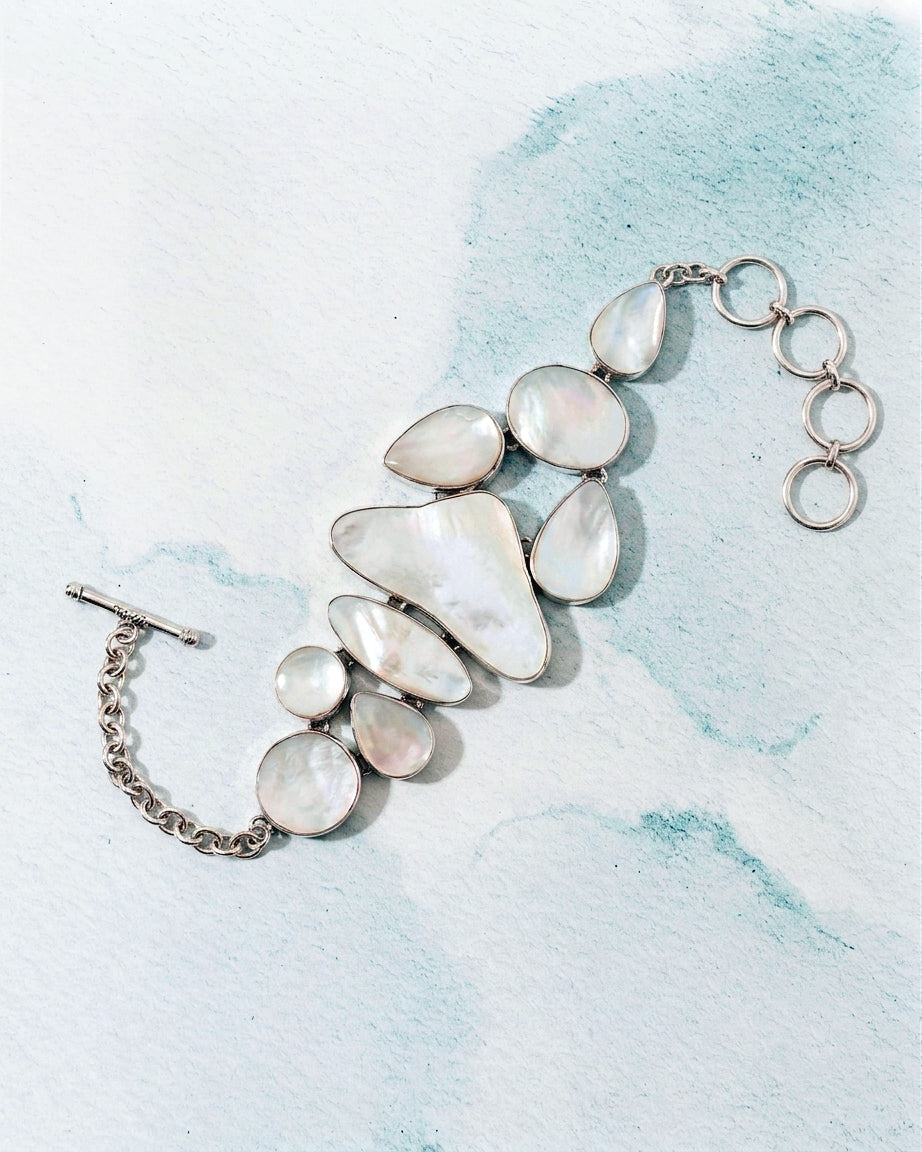 A silver-toned statement bracelet featuring irregular, organic-shaped white mother-of-pearl stones, displayed on a textured light blue and white background.