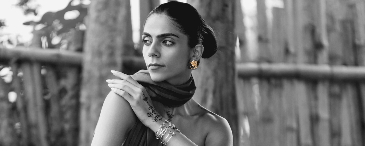 A black and white portrait of a woman wearing a dark scarf, earrings, and bracelets, posed in front of a rustic wooden fence wearing golden-green elephant shaped MALACHITE studs, with the rest of the image rendered in black and white.