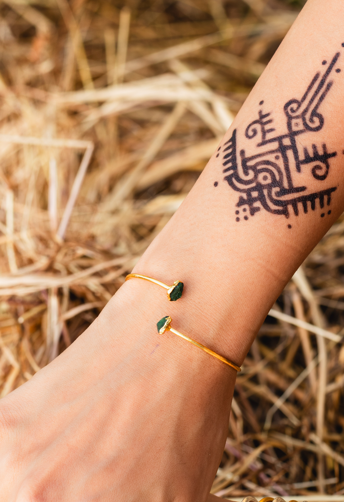 A close-up of a person's hands and wrists adorned with multiple delicate gold huggie bracelets, featuring small Green ONYX gemstone accents, set against a rustic background of dry grass.