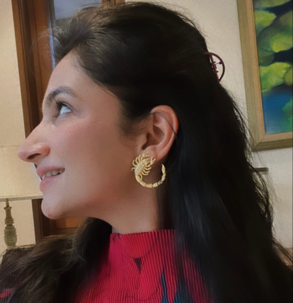 A person wearing a red top and scorpion earrings with a gold-plated scorpion design on their earlobes.