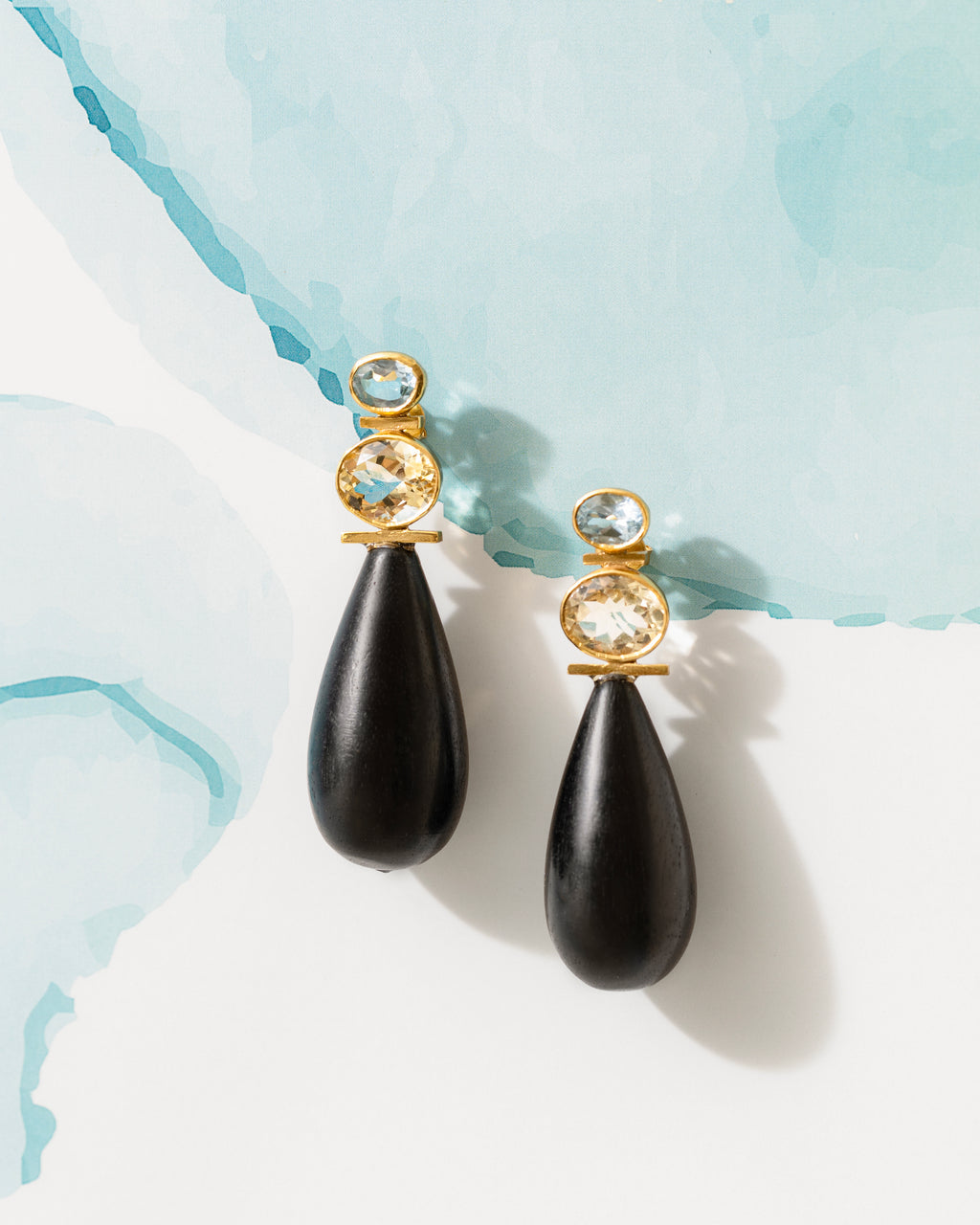 A pair of gold and black teardrop dangle earrings with clear gemstones, displayed on a white surface with a light blue watercolor background.