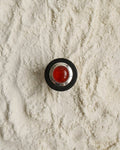 A round contemporary-style button with a red wooden CARNELIAN center and Eco-925 Sterling Silver-Ebony wood outer ring, placed on a textured background of white sand.