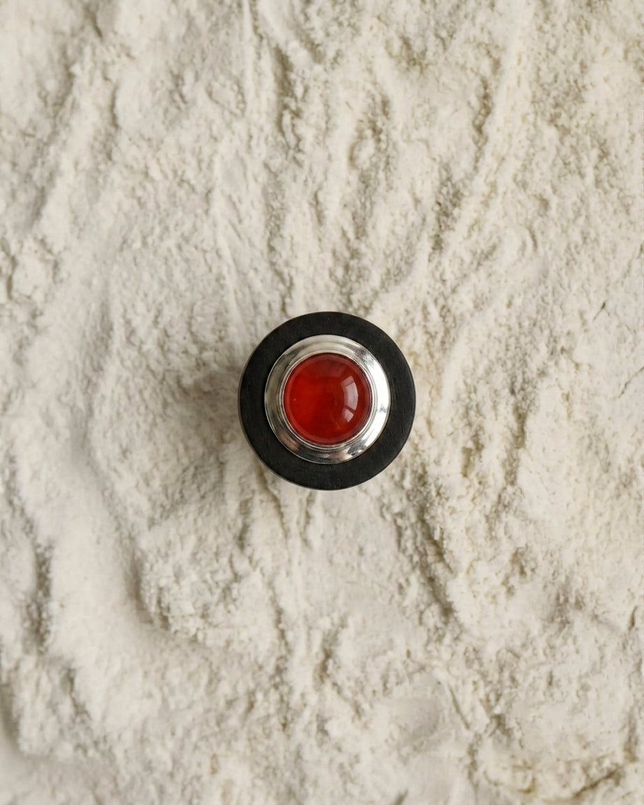 A round contemporary-style button with a red wooden CARNELIAN center and Eco-925 Sterling Silver-Ebony wood outer ring, placed on a textured background of white sand.