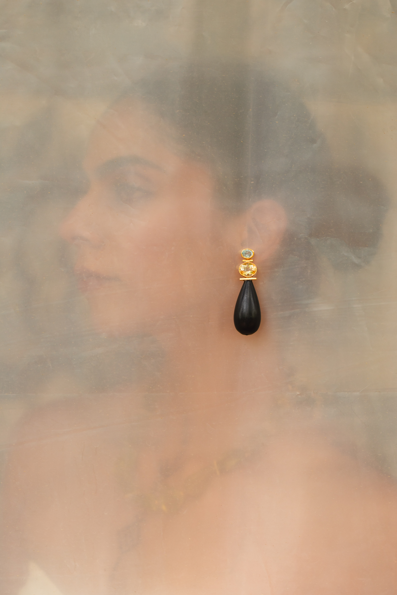 A single black teardrop dangle earring with a gold-toned top setting and a small clear stone, shown worn by a woman in a softly blurred, warm-toned portrait.