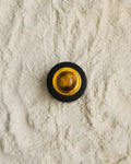 A round contemporary-style button or knob with a black Ebony wood outer ring, gold-toned inner ring, and a tiger-eye's gemstone in the center, placed on a textured off-white sand like surface.