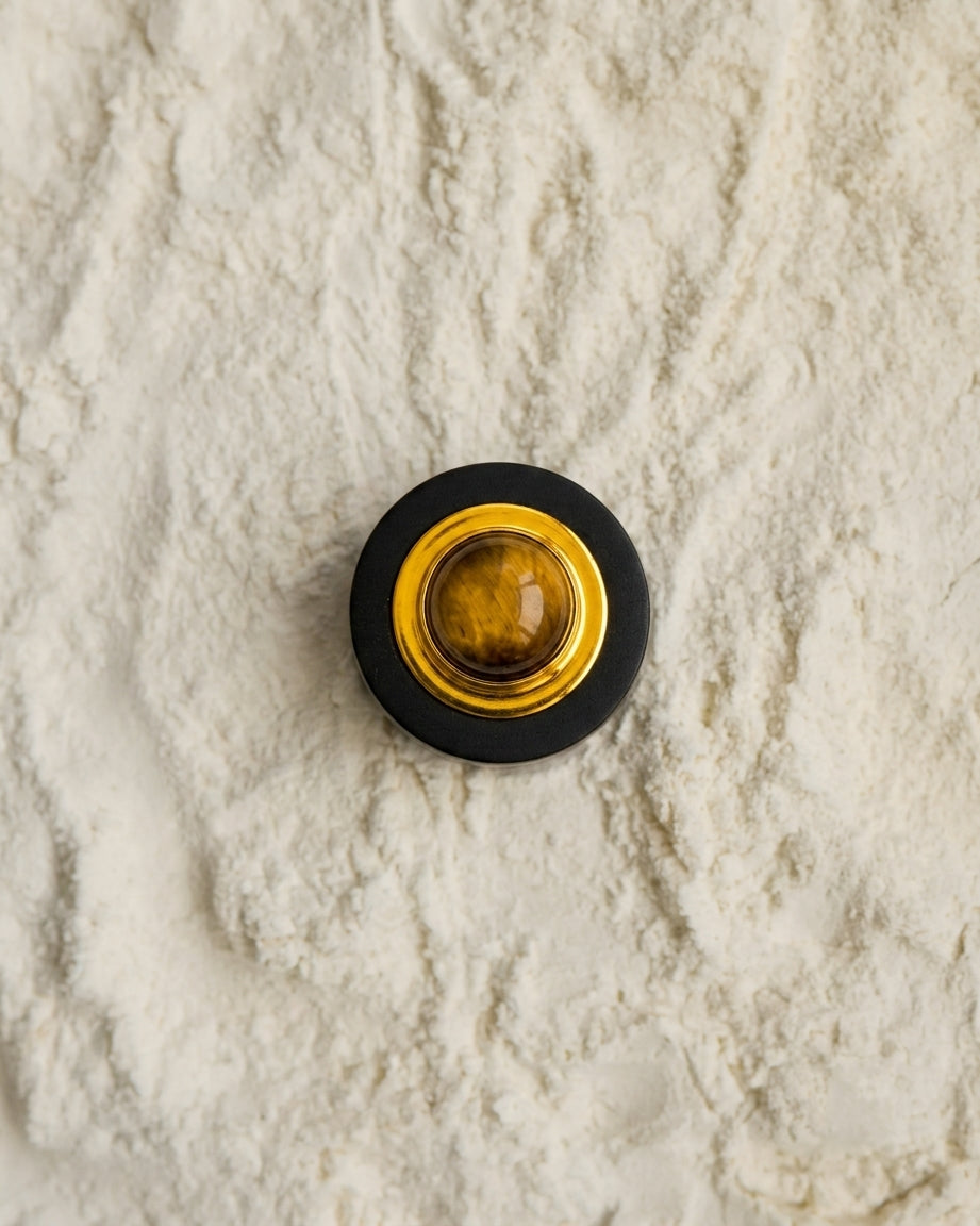 A round contemporary-style button or knob with a black Ebony wood outer ring, gold-toned inner ring, and a tiger-eye's gemstone in the center, placed on a textured off-white sand like surface.