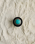 A round contemporary-style button or knob with a black Ebony wood outer ring, Eco-925 Sterling silver-toned inner ring, and a turquoise stone in the center, placed on a textured off-white sand like surface.