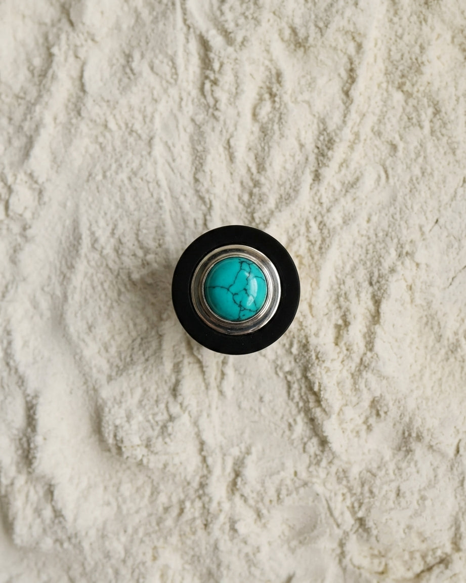 A round contemporary-style button or knob with a black Ebony wood outer ring, Eco-925 Sterling silver-toned inner ring, and a turquoise stone in the center, placed on a textured off-white sand like surface.
