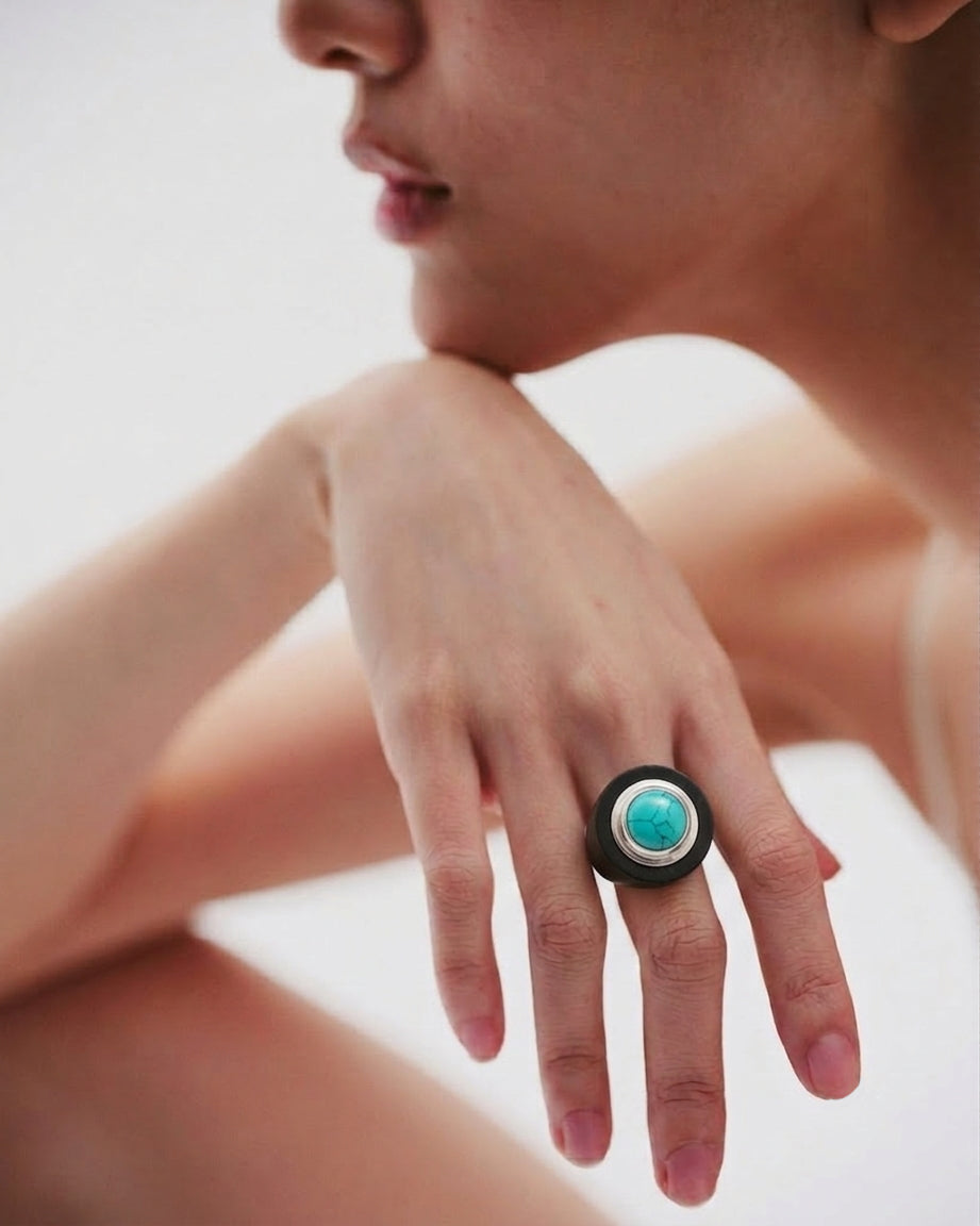A close-up lifestyle shot of a model wearing a large statement ring on her finger. The ring features a black ebony wood circular band with a central turquoise stone surrounded by a Eco-925 Sterling silver-toned bezel.